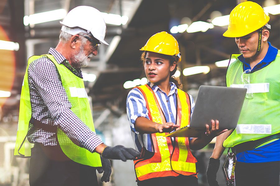 Industries - Warehouse Workers in PPE Discussing While Standing and the Middle Worker is Holding an Open Laptop