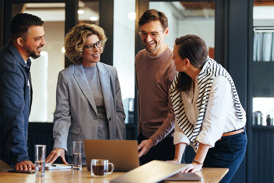 Insurance Products - Peers Smiling While Standing Around a Table with an Open Laptop as They Discuss in the Office