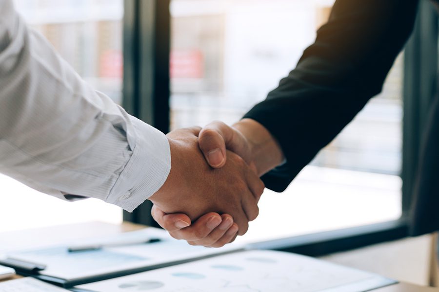 Partnership - Close Up of Two Professionals Shaking Hands in an Office While Standing Over a Table with Documents Next to a Window