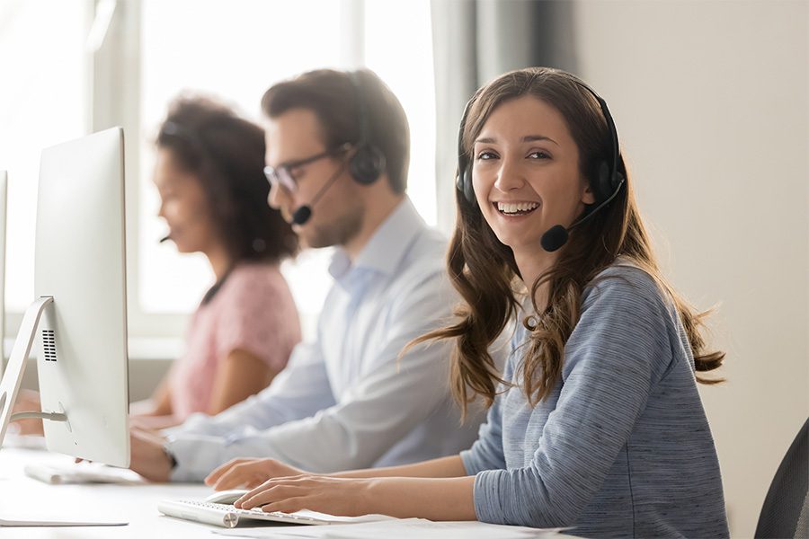 Client Center - Woman With Headset Smiling at Camera While Working on a Laptop and Her Coworkers are Behind Her Working as Well in the Office